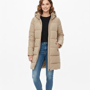 Può includere: Un lungo cappotto imbottito beige con cappuccio e chiusura a zip. Il cappotto è indossato sopra un top nero e jeans blu. La modella indossa scarpe da ginnastica bianche. Il cappotto ha un design trapuntato e tasche.