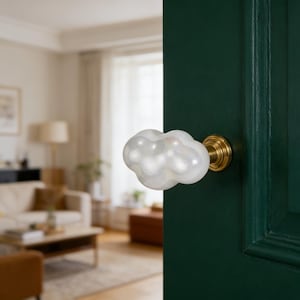 May include: A white, cloud-shaped glass door knob with a brass base, attached to a dark green door. The background shows a blurred living room scene.
