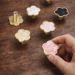 Peut inclure: Boutons de meubles en forme de fleur de différentes couleurs, dont blanc, rose et noir, avec des bases dorées. Un bouton est tenu par une main. Ces boutons décoratifs sont parfaits pour les meubles.