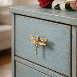 May include: A distressed light blue wooden bedside table with a gold dragonfly handle. The table has two drawers. A bouquet of flowers, including red roses and white lilies, sits on top. The table is approximately 60 cm tall.