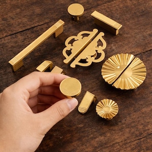 May include: A selection of gold-coloured cabinet hardware, including knobs, pulls, and decorative accents. The hardware is displayed on a dark wood surface, showcasing various designs and sizes. A hand is holding one of the knobs.