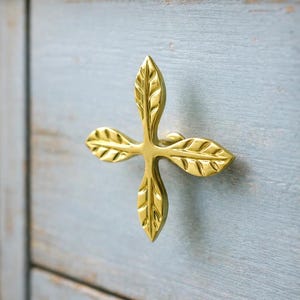 以下が含まれることがあります： 明るい青色の木製キャビネットに取り付けられた、金色の葉の形をした引き出しプル。 引き出しプルは、十字型に配置された 4 つの葉を持っています。 キャビネットは、塗装された、使い古された仕上げになっています。