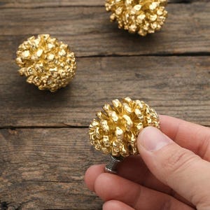 Può includere: Pomelli per mobili dorati e strutturati a forma di pigna. Un pomello è tenuto in mano, con altri due sparsi su una superficie di legno. I pomelli hanno una base a vite. Ferramenta decorativa.