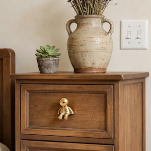 May include: A wooden bedside table with two drawers, each featuring a gold-coloured human figure knob. A vase of lavender and a small potted succulent sit on top. A light switch is visible on the wall.