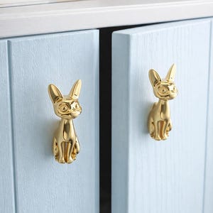 May include: Gold-colored cabinet pulls in the shape of a cat. The cat-shaped knobs are mounted on light blue cabinet doors. The cabinet doors are closed, and the knobs are centered on each door. The cabinet is part of a larger piece of furniture.