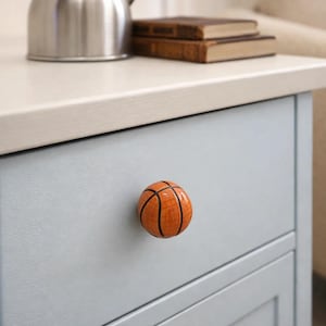 以下が含まれることがあります： バスケットボール型の引き出しノブが付いた水色のキャビネット。ノブはオレンジ色で黒い線があり、バスケットボールに似ています。ステンレス製のケトルと2冊の本がカウンタートップにあります。