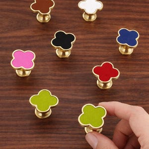 Könnte beinhalten: Eine Sammlung von kleeblattförmigen Schrankknöpfen in verschiedenen Farben, darunter Braun, Weiß, Schwarz, Blau, Rosa, Rot und Grün. Jeder Knopf hat einen goldfarbenen Sockel. Ein grüner Knopf wird von einer Hand gehalten.