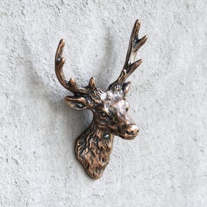 Puede incluir: Un gancho de pared de color bronce con forma de cabeza de ciervo, con astas detalladas y pelaje texturizado. La escultura presenta un acabado metálico con reflejos oscuros en las hendiduras, capturando los rasgos faciales realistas del animal.
