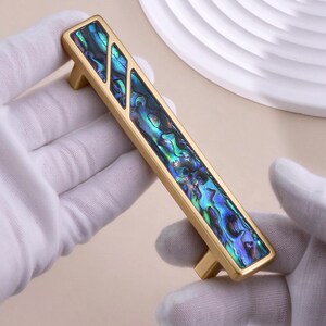 Puede incluir: Un tirador de mueble rectangular con acabado en oro mate y una tira incrustada de concha de abulón iridiscente. La concha presenta patrones en espiral con tonos azules, verdes y morados. El tirador tiene dos puntos de montaje en la base.