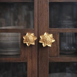 Może przedstawiać: Zbliżenie na ciemną drewnianą szafkę ze szklanymi drzwiami. Drzwi szafki mają złote gałki w kształcie słoneczników. Wnętrze szafki jest widoczne, z półkami i sugestią przechowywanych w niej przedmiotów.