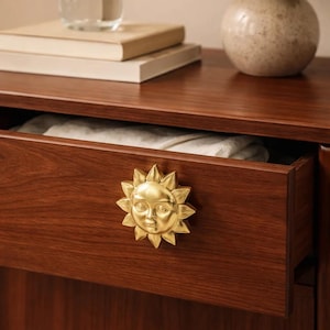 Puede incluir: Una cómoda de madera con un cajón que presenta un pomo dorado en forma de sol. El cajón está abierto, revelando un paño blanco doblado. Un jarrón con flores, libros y un vaso de agua están encima.