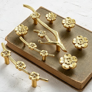 May include: A collection of gold-colored cabinet hardware, including knobs and pulls, arranged on a vintage book. The hardware features a floral design, with each piece showcasing detailed flower shapes. The pulls are branch-like, with multiple flower accents.