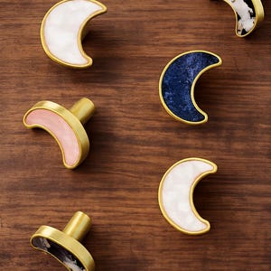 May include: Decorative crescent moon cabinet knobs in various colors. The knobs have a gold-colored base and a crescent-shaped top in white, pink, navy blue, and black and white marbled patterns. These knobs are ideal for adding a unique touch to furniture.
