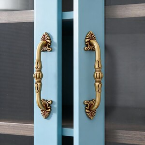 May include: A pair of antique brass door handles with decorative floral detailing. The handles are fixed to a light blue cabinet door. The handles are curved and are likely used for opening and closing a cabinet or door.
