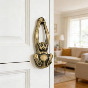 Può includere: Un battente per porta a forma di scimmia color ottone. La scimmia è raffigurata appesa con le braccia alzate, che tiene una barra. Il battente è montato su una porta bianca, con uno scorcio di un soggiorno sullo sfondo.