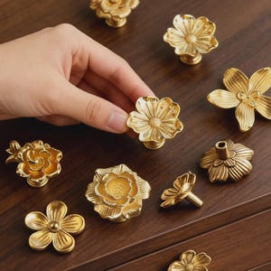 以下が含まれることがあります： さまざまなデザインの、金色の花の形をしたキャビネットノブ。ノブは金属製で、詳細な花の模様があります。いくつかのノブは手で持たれており、さまざまなスタイルが紹介されています。