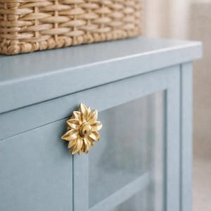 Op de afbeelding: Een lichtblauwe kast met een gouden handvat in de vorm van een zonnebloem. De kast heeft glazen deuren en een geweven mand bovenop. De kast is geschilderd in een zachte, gedempte blauwe kleur.