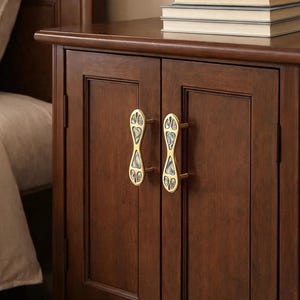May include: A dark brown wooden nightstand with two doors and decorative gold-colored handles. The handles have an hourglass shape with iridescent inlays. A stack of books sits on top of the nightstand, next to a bed.