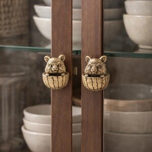 Könnte beinhalten: Nahaufnahme eines Holzschranks mit zwei messingfarbenen Schrankgriffen. Jeder Griff zeigt einen detaillierten, geformten Bärenkopf, der aus einer dekorativen Schale ragt. Die Schranktüren sind dunkelbraun.