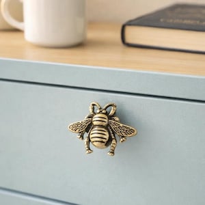May include: A brass-colored bee-shaped drawer pull is mounted on a light blue drawer. The bee has detailed wings and body stripes. A white mug and a book are visible in the background, suggesting a home decor theme.