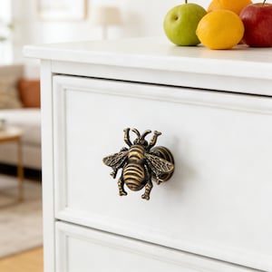 Könnte beinhalten: Dekorativer Schubladengriff in Form einer Biene aus Bronze, montiert an einem weißen Schrank. Die Biene hat detaillierte Flügel und Körperstreifen. Der Schrank hat ein klassisches Design, mit Obst oben.
