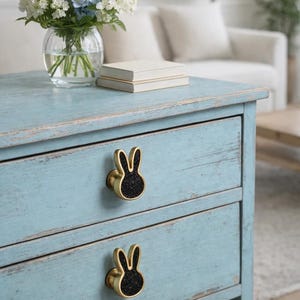 Peut inclure: Une commode en bois peinte en bleu clair avec deux tiroirs. Chaque tiroir est orné d'un bouton doré en forme de lapin avec un centre noir pailleté. Un vase de fleurs et deux livres sont posés dessus.
