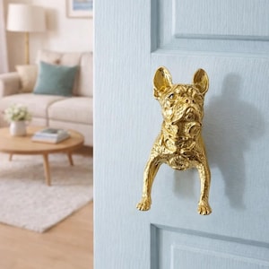 May include: A gold-colored door handle shaped like a French Bulldog. The dog-shaped handle is mounted on a light blue door. The handle is a decorative accent for home decor.