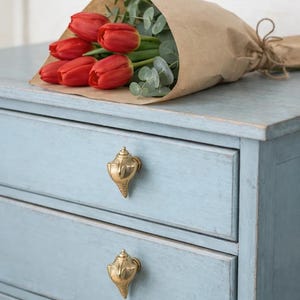 Könnte beinhalten: Ein Strauß roter Tulpen und Eukalyptus, eingewickelt in braunes Papier, liegt auf einer hellblauen Kommode. Die Kommode hat zwei Schubladen mit goldenen Muschelknöpfen. Der Stil ist rustikal und charmant.