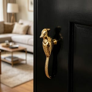 May include: A gold-coloured bird-shaped door handle fixed to a black door. The handle is an ornate piece with detailed features, adding a touch of elegance to the door's design. The handle's design is reminiscent of a bird.