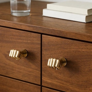 May include: Close-up of a wooden dresser with two brass fist-shaped drawer pulls. A glass of water and two books are on top of the dresser. The wood has a warm, brown tone.