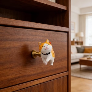 Op de afbeelding: Een grillige kastknop met een lachende oranje en witte kat met een zwarte strik. De katvormige knop is gemonteerd op een messingkleurige basis en bevestigd aan een donkerbruine houten lade. De lade maakt deel uit van een houten kast.