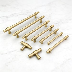 Puede incluir: Colección de tiradores y pomos dorados para armarios de varios tamaños. La ferretería incluye tiradores de barra y pomos en forma de T. Los artículos están dispuestos sobre una superficie de mármol blanco, mostrando su diseño y acabado.