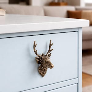 Peut inclure: Poignée de tiroir en forme de tête de cerf en bronze, fixée sur une armoire bleu clair. La tête de cerf présente des bois et des traits du visage détaillés. L'armoire fait partie d'un meuble plus grand.