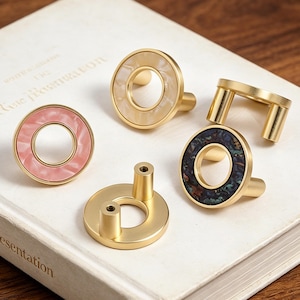 May include: A collection of decorative cabinet knobs and pulls in various designs. The knobs feature gold-toned metal with circular accents in pink, cream, and a dark, speckled pattern. One pull is a simple gold bar design.