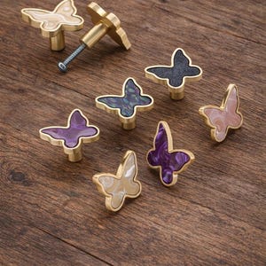 Peut inclure: Ensemble de boutons de meubles en forme de papillon, de couleurs variées : violet, rose et noir, avec des éléments dorés. Les boutons sont disposés sur une surface en bois.