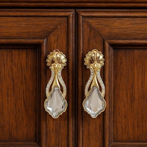 Puede incluir: Primer plano de dos tiradores dorados ornamentados con detalles de cristal transparente. Los tiradores están montados en una puerta de armario de madera marrón oscuro. Los tiradores tienen un diseño floral en la parte superior y un cristal en forma de lágrima.