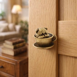 Könnte beinhalten: Bronzefarbener Türknauf in Katzenform, der eine schlafende Katze in einer Teetasse zeigt. Der Knauf ist an einer hellen Holztür befestigt. Die Teetasse steht auf einer passenden Untertasse und wirkt dekorativ.