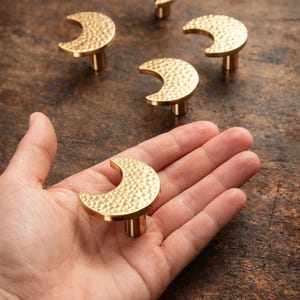 Puede incluir: Pomos dorados para armarios con forma de media luna y superficie texturizada. Los pomos se muestran sobre una superficie de madera, con un pomo en la mano. Estos pomos decorativos son adecuados para muebles.