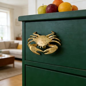 Puede incluir: Una cómoda verde oscuro con un tirador dorado en forma de cangrejo. La cómoda está coronada con varias frutas, incluyendo manzanas y naranjas. El tirador de cangrejo es un acento detallado de temática náutica.