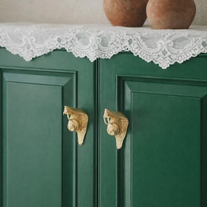 May include: A dark green cabinet with two gold-colored snake-shaped handles. Two terracotta pots with handles sit on top of the cabinet, which is covered with a white lace doily.