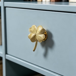 May include: A gold-toned metal cabinet knob shaped like a four-leaf clover with a textured, ribbed surface on each leaf. The clover knob is mounted on a flat, circular base and attached to a smooth, matte light blue drawer front surface.