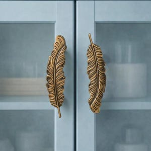 May include: Two bronze-colored feather-shaped cabinet pulls mounted on a light blue cabinet. The cabinet doors have glass panels, and the background shows shelves with white objects. Woven baskets sit above the cabinet.