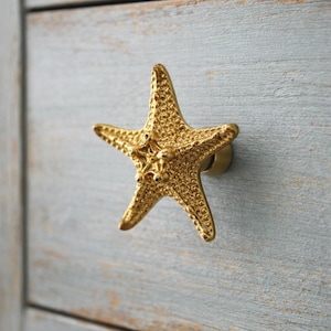 以下が含まれることがあります： 金色のヒトデ型キャビネットノブ。明るい青色の木製キャビネットドアに取り付けられています。ヒトデはテクスチャ加工された表面を持ち、海岸や航海をテーマにした家具の装飾的なアクセントです。