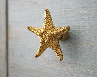 Peut inclure: Un bouton de meuble en forme d'étoile de mer de couleur dorée. Le bouton est monté sur une porte d'armoire en bois bleu clair. L'étoile de mer a une surface texturée et est un élément décoratif pour les meubles sur le thème côtier ou nautique.