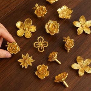May include: A collection of gold-colored floral cabinet knobs and pulls in various designs, including roses, daisies, and abstract flower shapes. The hardware is displayed on a dark wood surface.