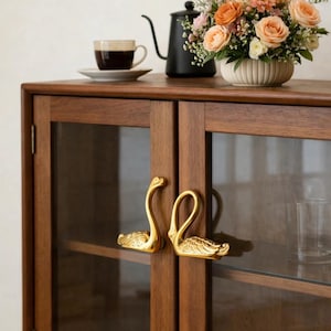 May include: A wooden cabinet with glass doors features gold swan-shaped handles. The cabinet is topped with a coffee cup, a black kettle, and a floral arrangement of peach and pink roses in a white vase.