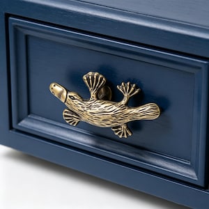 Op de afbeelding: Close-up van een marineblauwe lade met een decoratieve messing platypus-vormige handgreep. De handgreep is gedetailleerd met getextureerde lijnen en heeft een kleine snavel en zwemvoeten. De lade heeft een verzonken paneelontwerp.
