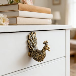 Può includere: Un pomello per cassetto a forma di pavone in ottone dettagliato è montato su un cassetto bianco. Il pavone è rivolto verso destra con le penne della coda spiegate. Il cassetto fa parte di un comò bianco. Libri e fiori sullo sfondo.