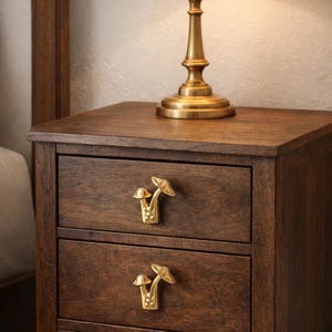Könnte beinhalten: Ein dunkelbrauner Nachttisch mit zwei Schubladen, die jeweils mit goldenen, pilzförmigen Griffen verziert sind. Auf der Oberseite steht eine Messinglampe mit cremefarbenem Schirm. Teil eines Schlafzimmermöbels.