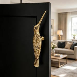 May include: A gold-colored door handle in the shape of a hummingbird, mounted on a black door. The handle is detailed with textured patterns and a long beak. The background shows a living room with a sofa and a coffee table.